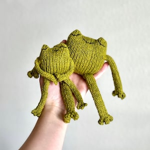 May include: Two green knitted frog plushies, one larger than the other, with smiling faces. The frogs are sitting on a hand.