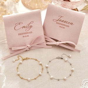 May include: Two light pink velvet jewellery pouches with ribbon bows, each printed with a name, the word BRIDESMAID, and the date 10.11.26. Below the pouches are two delicate bracelets, one gold-toned and one silver-toned, both featuring small white pearls.