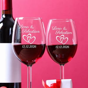 Può includere: Due calici da vino trasparenti riempiti di vino rosso, ciascuno inciso con i nomi Lena & Sebastian, due cuori intrecciati e la data 12.12.2026. Sono presenti una bottiglia di vino, un piccolo cuore rosso e una scatola regalo bianca con nastro.