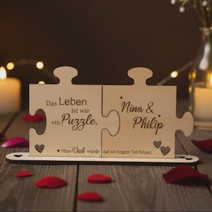Può includere: Un espositore in legno chiaro composto da due pezzi di puzzle a incastro. Il testo recita: Das Leben ist wie ein Puzzle. Nina & Philip. Ohne Dich würde das wichtigste Teil fehlen. Sono presenti piccoli intagli a forma di cuore.