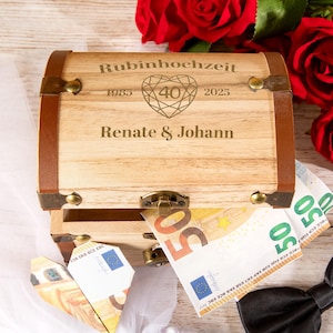 Könnte beinhalten: Eine Holzkiste mit braunen Lederbesätzen und Metallbeschlägen. Die Kiste ist mit den Worten "Rubinhochzeit", "1985", "40", "2025" und "Renate & Johann" graviert. Fünfzig-Euro-Banknoten sind im Inneren der Kiste sichtbar.