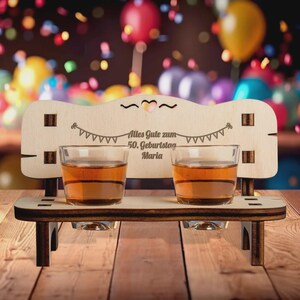 Könnte beinhalten: Ein Holz-Shotglashalter in Form einer Bank mit zwei Shotgläsern, die mit bernsteinfarbener Flüssigkeit gefüllt sind. Der Halter hat dekorative Ausschnitte und den Text "Alles Gute zum 50. Geburtstag Maria". Bunte Konfetti und Luftballons sind im Hintergrund.