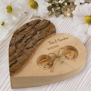 Puede incluir: Un portaanillos de madera en forma de corazón con dos anillos de oro. El soporte tiene una sección de corteza y está grabado con "Tim & Sandra" y la fecha 20.08.2020. Un lazo de cuerda asegura los anillos.