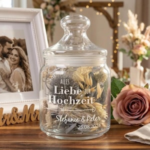 Op de afbeelding: Een heldere glazen pot met een deksel, gevuld met gedroogde bloemen en bladeren. De pot heeft witte tekst met de tekst "Alles Liebe zur Hochzeit" en de namen "Stefanie & Peter" met de datum "25.08.2024". Een ingelijste foto en een roos staan op de achtergrond.