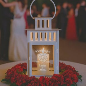 Op de afbeelding: Een witte lantaarn met een handvat, met een brandende kaars. De kaars is gegraveerd met de woorden "Rubinhochzeit", "40" en namen. De lantaarn is omgeven door een krans van rode bloemen, met mensen op de achtergrond.