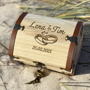 Op de afbeelding: Een houten schatkist met bruine leren accenten en metalen beslag. De kist is gegraveerd met de namen "Lena & Tim", trouwringen en de datum "25.02.2025". Een sleutel zit in het slot.