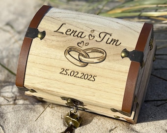 Könnte beinhalten: Eine hölzerne Schatzkiste mit braunen Lederakzenten und Metallbeschlägen. Die Kiste ist mit den Namen "Lena & Tim", verschlungenen Eheringen und dem Datum "25.02.2025" graviert. Ein Schlüssel steckt im Schloss.