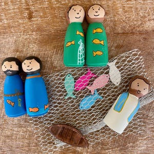Puede incluir: Muñecos de madera pintados que representan figuras bíblicas. Los muñecos visten túnicas verdes y azules, con motivos de peces y redes. También hay un pequeño bote de madera y peces de colores, colocados sobre una red de pesca.