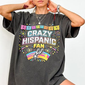 May include: Dark gray t-shirt with the text "CRAZY HISPANIC FAN" in colorful lettering. The design includes fireworks, a rainbow, and a stylized logo. The shirt has short sleeves and a relaxed fit.