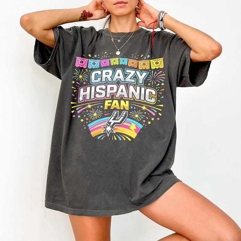 May include: Dark gray t-shirt with the text "CRAZY HISPANIC FAN" in colorful lettering. The design includes fireworks, a rainbow, and a stylized logo. The shirt has short sleeves and a relaxed fit.