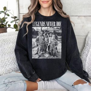 May include: Black crewneck sweatshirt with a grayscale image of a group of people. The image is centered on the front of the sweatshirt and has the text "LEGENDS NEVER DIE" above it in white block letters.