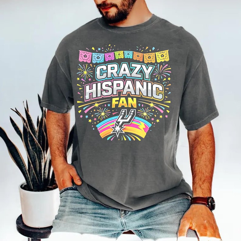 May include: Dark gray t-shirt with the text "CRAZY HISPANIC FAN" in a colorful design. The shirt features a festive banner and fireworks, with a basketball team logo at the bottom. The shirt is a casual wear item.