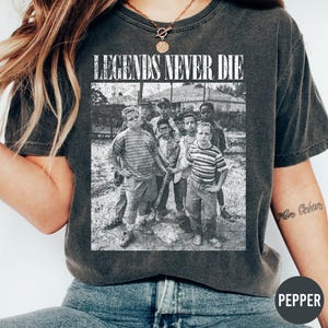 May include: Dark gray t-shirt featuring a black and white image of a group of boys. The text "LEGENDS NEVER DIE" is printed above the image. The shirt is a vintage style.