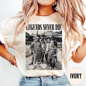 May include: Ivory-colored sweatshirt featuring a black and white graphic of a group of boys with the text "LEGENDS NEVER DIE" above the image. The sweatshirt has a crew neck and long sleeves. The graphic is a vintage-style photograph.