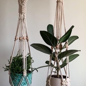 May include: Two macrame plant hangers with wooden beads. One holds a teal ceramic pot with a green plant, the other a white pot with a leafy green plant. The hangers are suspended from wooden rings.