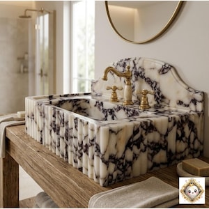 May include: A marble bathroom sink with a white and black veined pattern. The sink has a gold faucet and is set on a wooden vanity. The bathroom has a neutral color palette with a shower and a potted olive tree.