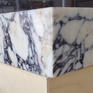 May include: A rectangular sink basin crafted from white marble featuring prominent, dark purple and black veining throughout the stone surface. The basin has a smooth, polished finish and is positioned on a light-colored wooden support structure.