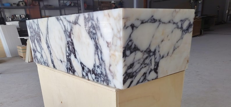 May include: A rectangular sink basin crafted from white marble featuring prominent, dark purple and black veining throughout the stone surface. The basin has a smooth, polished finish and is positioned on a light-colored wooden support structure.