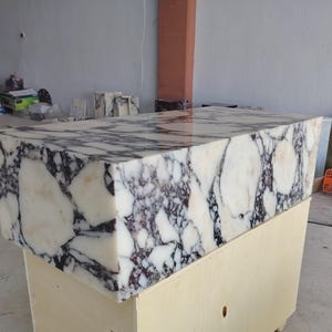 May include: A rectangular block of polished white marble featuring prominent, dark purple and black veining throughout its surface. The stone has a smooth, reflective finish and is positioned atop a light-colored wooden crate base.