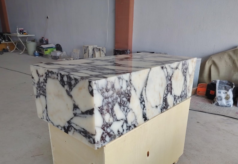 May include: A rectangular block of polished white marble featuring prominent, dark purple and black veining throughout its surface. The stone has a smooth, reflective finish and is positioned atop a light-colored wooden crate base.