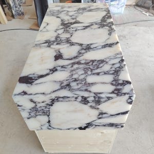 May include: A rectangular table with a polished white marble top featuring prominent dark purple and black veining. The table is supported by a sturdy, light-colored wooden base, showcasing a high-contrast stone pattern across the flat surface.