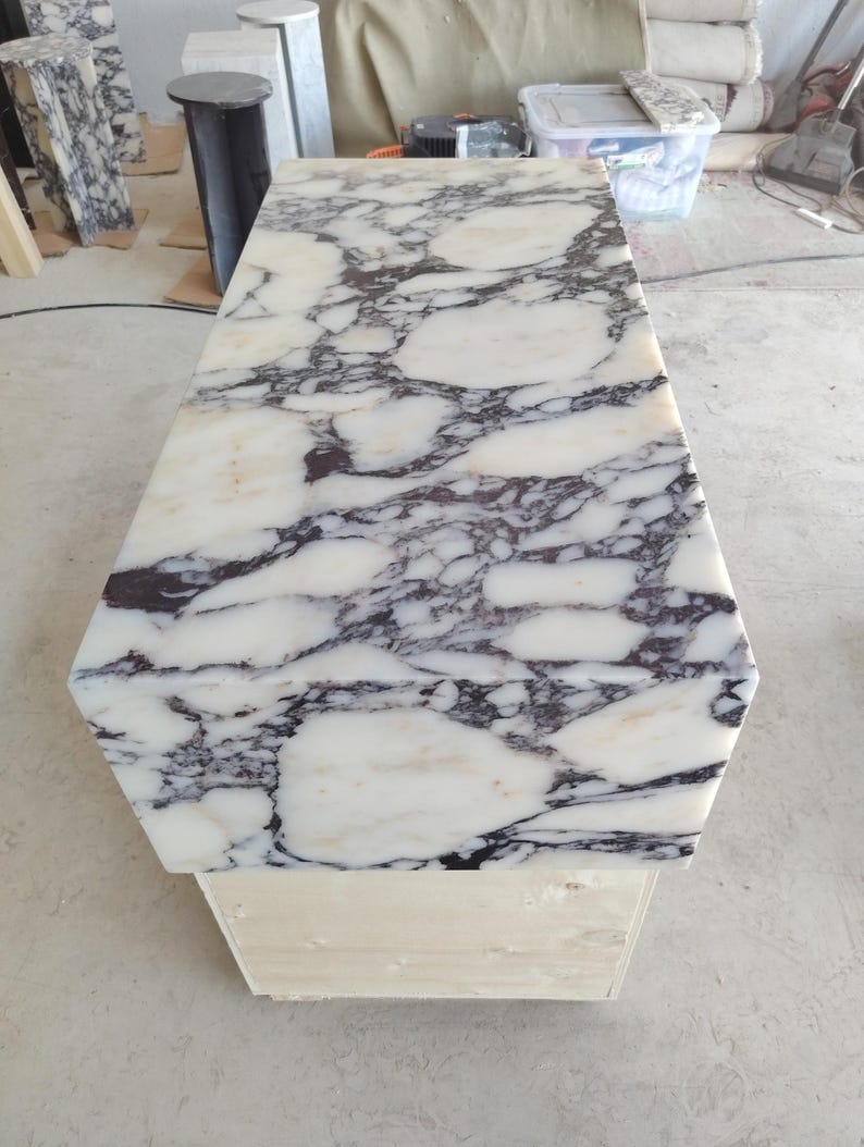May include: A rectangular table with a polished white marble top featuring prominent dark purple and black veining. The table is supported by a sturdy, light-colored wooden base, showcasing a high-contrast stone pattern across the flat surface.