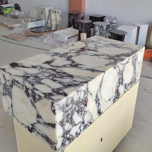 May include: A rectangular block of white marble featuring prominent, irregular dark purple and black veining throughout its surface. The stone block has a polished finish and is positioned atop a light-colored wooden base in a workshop setting.