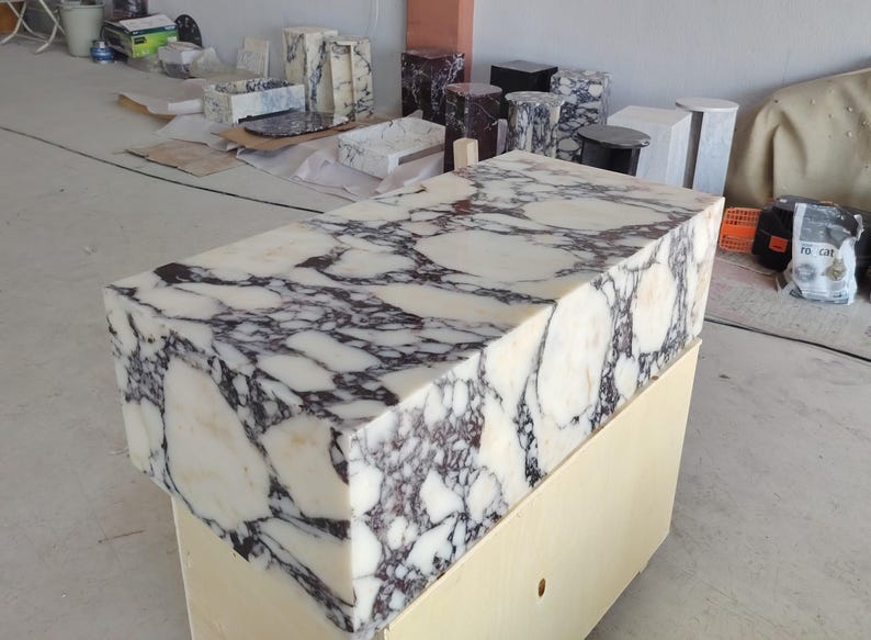 May include: A rectangular block of white marble featuring prominent, irregular dark purple and black veining throughout its surface. The stone block has a polished finish and is positioned atop a light-colored wooden base in a workshop setting.