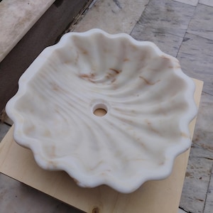May include: A white marble sink with a scalloped edge and a central drain hole. The sink has subtle brown veining throughout, adding a natural aesthetic. The sink is placed on a light-colored wooden surface.