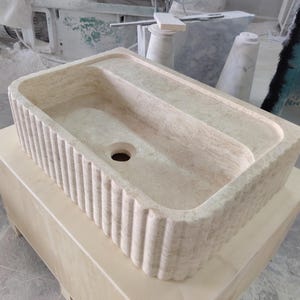 May include: A rectangular, beige stone sink with a ribbed exterior. The sink has a central drain hole and a raised, flat area on one side. It sits on a light-coloured wooden cabinet. The sink's design suggests a focus on natural materials and a minimalist aesthetic.