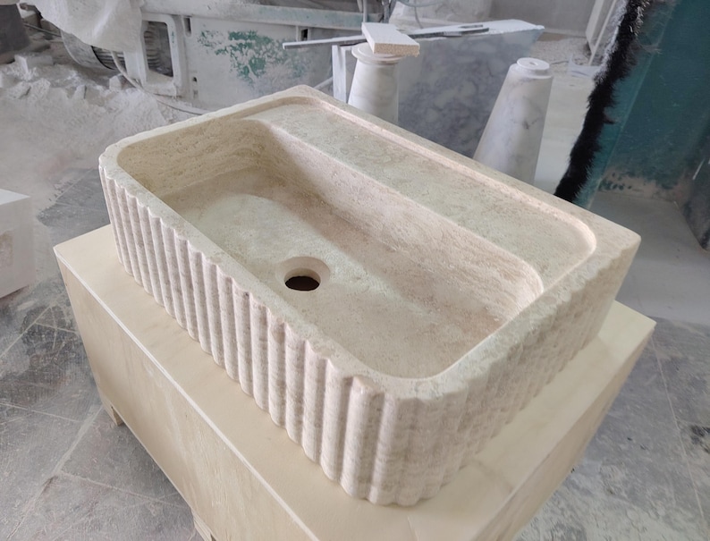 May include: A rectangular, beige stone sink with a ribbed exterior. The sink has a central drain hole and a raised, flat area on one side. It sits on a light-colored wooden cabinet. The sink's design suggests a focus on natural materials and a minimalist aesthetic.