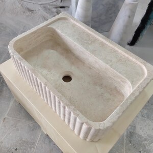 May include: A rectangular, off-white stone sink with a fluted front edge. The sink has a built-in ledge on one side and a drain hole in the center. The sink is sitting on a light-colored wooden box.