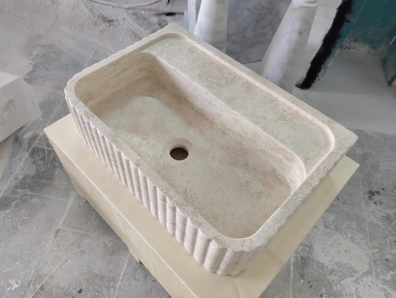 May include: A rectangular, off-white stone sink with a fluted front edge. The sink has a built-in ledge on one side and a drain hole in the center. The sink is sitting on a light-colored wooden box.