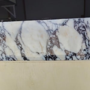 May include: A rectangular slab of white marble featuring prominent, dark purple and black veining throughout the surface. The stone displays a polished finish with natural color variations, including subtle hints of tan and brown within the patterns.