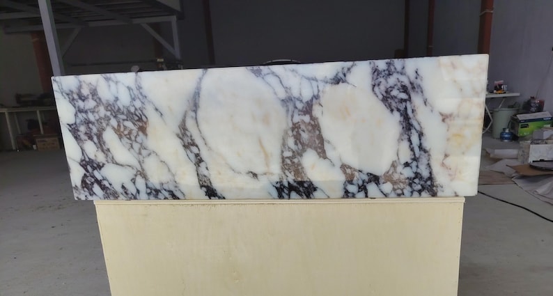 May include: A rectangular slab of white marble featuring prominent, dark purple and black veining throughout the surface. The stone displays a polished finish with natural color variations, including subtle hints of tan and brown within the patterns.