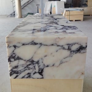 May include: A rectangular block of polished white marble featuring prominent, dark charcoal-colored veining throughout the surface. The stone slab is mounted on a plain, light-colored wooden base, showcasing a smooth, reflective finish on the top.