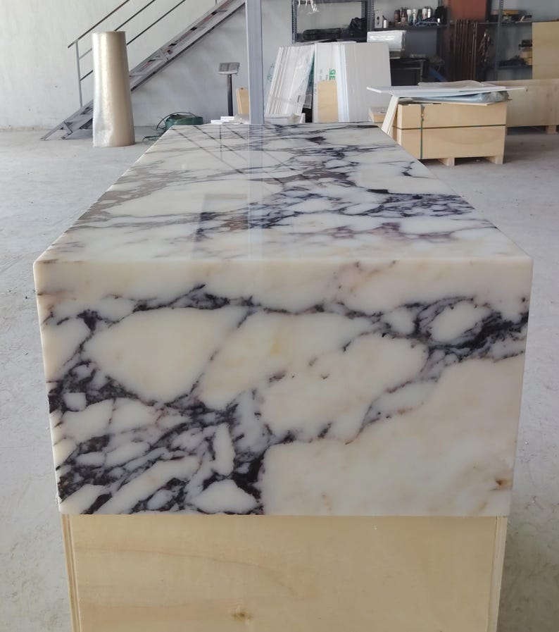 May include: A rectangular block of polished white marble featuring prominent, dark charcoal-colored veining throughout the surface. The stone slab is mounted on a plain, light-colored wooden base, showcasing a smooth, reflective finish on the top.