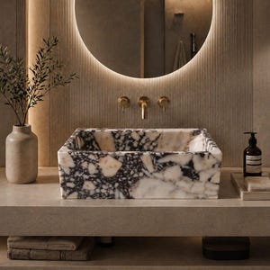 May include: A rectangular marble sink with a black and white pattern sits on a light-colored countertop. The bathroom features a round mirror with a warm light, gold fixtures, and a vase with greenery.