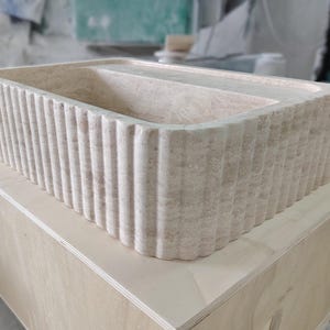 May include: A rectangular, beige stone sink with a fluted exterior. The sink is placed on a light-colored wooden box. The sink's interior is smooth and divided into two sections.