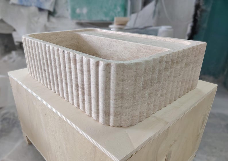 May include: A rectangular, beige stone sink with a fluted exterior. The sink is placed on a light-colored wooden box. The sink's interior is smooth and divided into two sections.