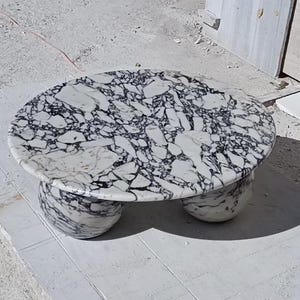 May include: A round coffee table crafted from white marble with prominent black veining. The tabletop features a polished surface with a complex, fragmented pattern, supported by three rounded, bulbous legs made of the same matching stone material.