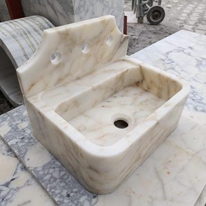 May include: A rectangular marble sink with a backsplash featuring three circular holes. The sink basin has a rounded front edge and a central drain hole. The marble has a cream and brown veined pattern.