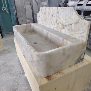 May include: A rectangular marble sink with a backsplash. The sink is a light beige colour with darker veining. It has a rectangular basin with a drain hole and is set on a wooden crate.