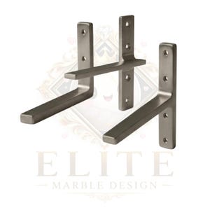May include: Three brushed metal shelf brackets. Each bracket has a vertical plate with pre-drilled holes for mounting and a horizontal arm. The brackets are a neutral grey colour. The text "ELITE MARBLE DESIGN" is at the bottom.