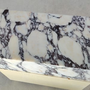 May include: A rectangular block with a polished white surface featuring prominent dark purple and black veining patterns. The top section is made of stone, while the base is a solid, smooth, light cream-colored material with a matte finish.