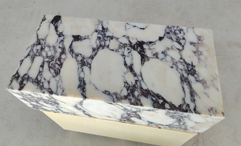 May include: A rectangular block with a polished white surface featuring prominent dark purple and black veining patterns. The top section is made of stone, while the base is a solid, smooth, light cream-colored material with a matte finish.