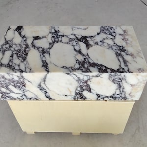 May include: A rectangular side table featuring a thick, polished white marble top with prominent dark purple and black veining. The base of the table is constructed from light-colored, smooth wood with a simple, box-like design and small feet.