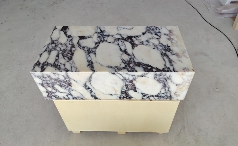 May include: A rectangular side table featuring a thick, polished white marble top with prominent dark purple and black veining. The base of the table is constructed from light-colored, smooth wood with a simple, box-like design and small feet.