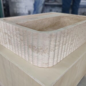 May include: A rectangular, beige stone sink with a fluted exterior. The sink is set on a light-colored wooden box. The sink's interior is smooth, while the exterior features vertical, ridged detailing. The overall design is minimalist and natural.