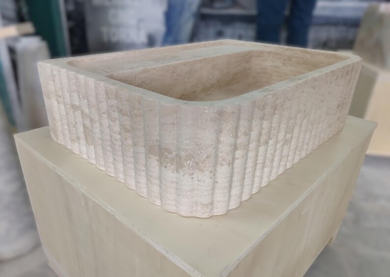 May include: A rectangular, beige stone sink with a fluted exterior. The sink is set on a light-colored wooden box. The sink's interior is smooth, while the exterior features vertical, ridged detailing. The overall design is minimalist and natural.